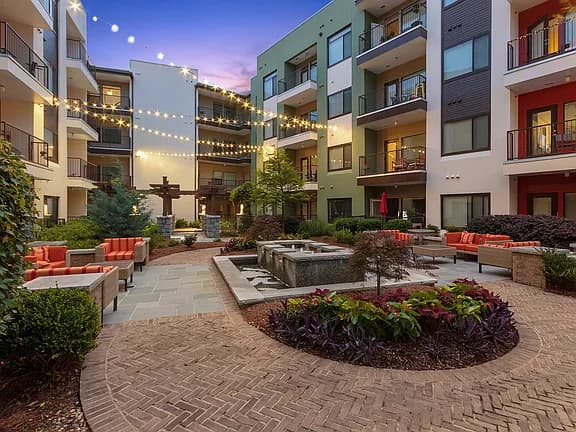 Courtyard at dusk with central fire pit, red seating areas, landscaped beds, and multi-story residential buildings with lit windows.