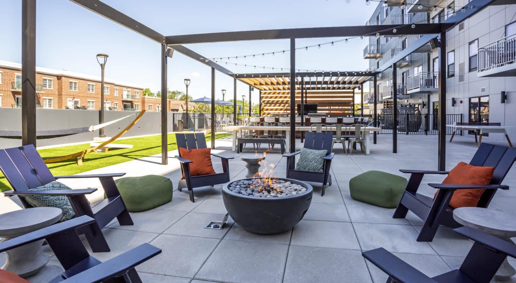 Rooftop terrace with central fire pit, Adirondack chairs with navy and orange cushions, green ottomans, pergolas with string lights, and multi-story residential buildings.