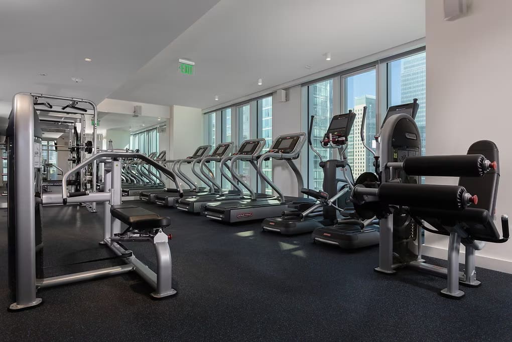 Modern gym with treadmills, ellipticals, weight machines, and dumbbells; floor-to-ceiling windows with downtown San Francisco skyline views.