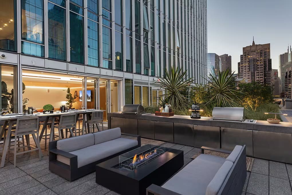 Rooftop terrace with gray modular seating, linear fireplace, potted plants, and San Francisco skyline backdrop.