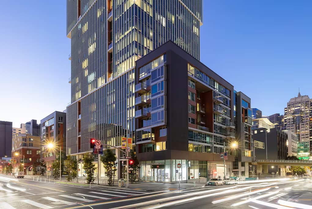 Modern mixed-use building with dark facade and blue-framed windows at street corner in downtown San Francisco, surrounded by taller office towers and city streets.