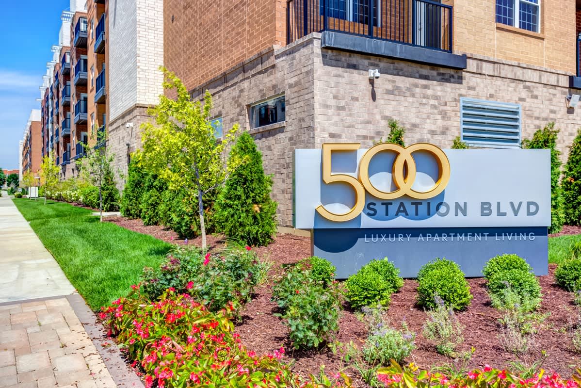 Exterior signage for 500 Station apartment complex with brick building facade, green shrubs, and red flowering plants along the entrance.