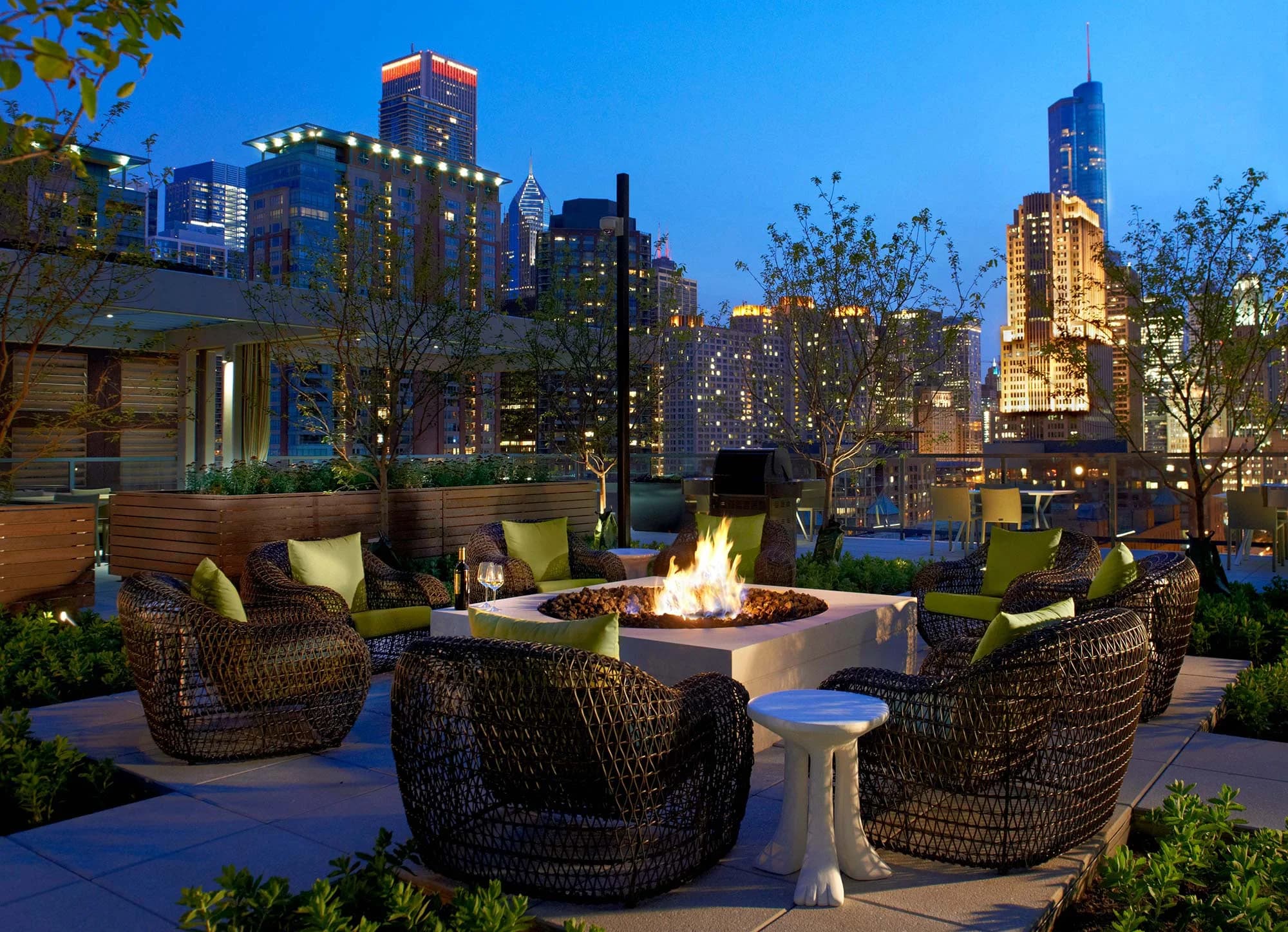 Rooftop terrace with woven lounge chairs arranged around a central fire pit, illuminated cityscape in background.