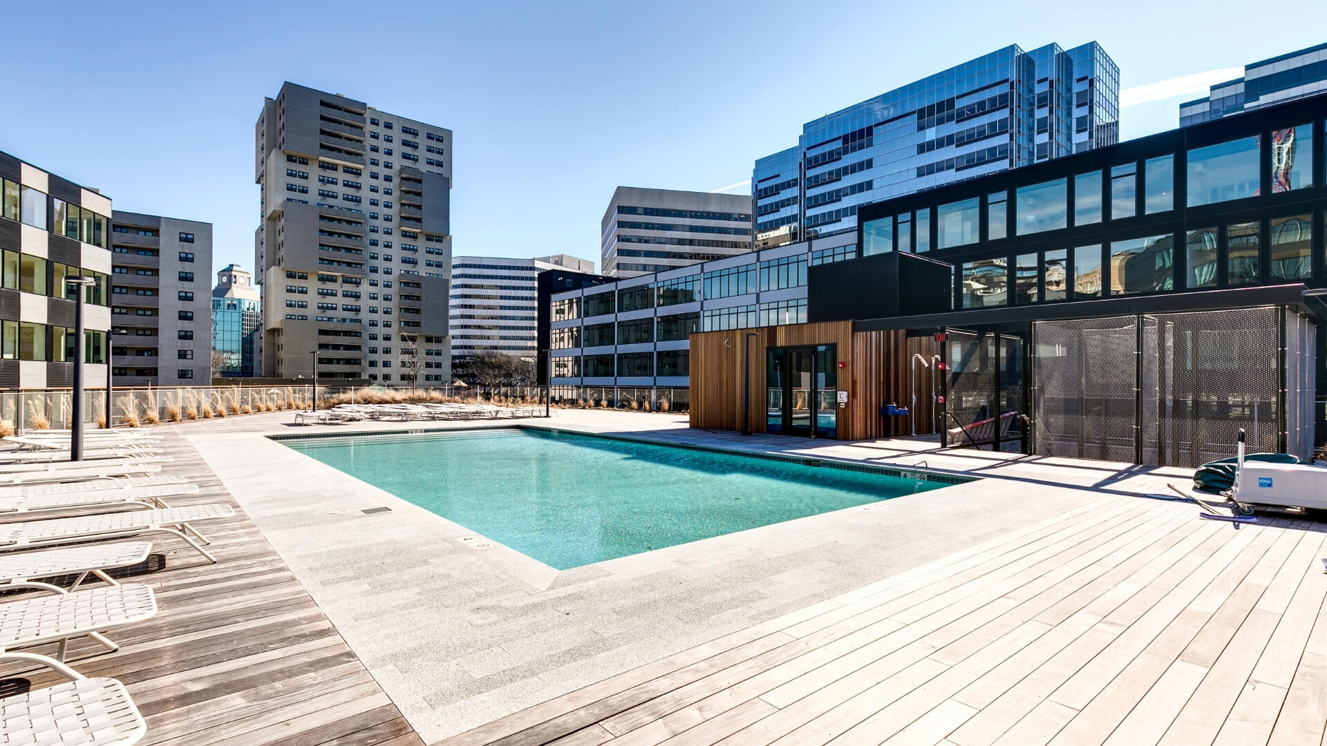 Rooftop pool with turquoise water, wooden deck lounge chairs, and modern high-rise buildings surrounding the amenity area.