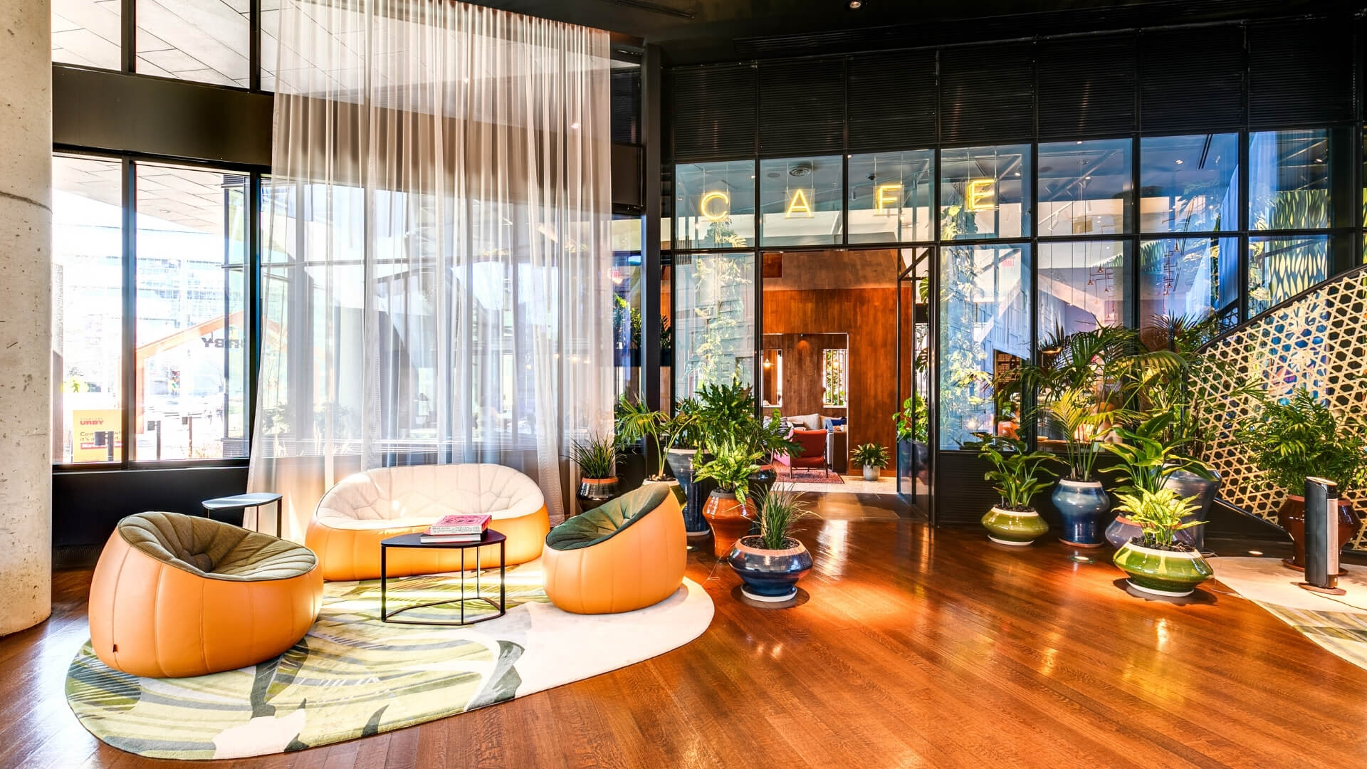 Modern lounge with orange pod chairs, small black table, potted plants, and large windows with city views.