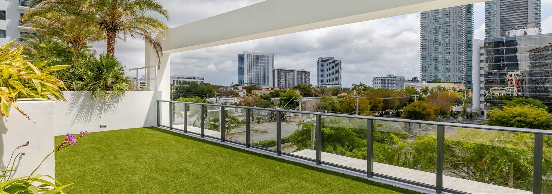 Balcony with green artificial turf, potted palm plants, glass railing, and panoramic view of Miami downtown high-rises and tree-lined streets.