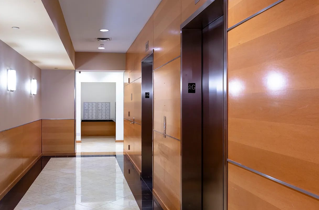 Building elevator lobby with wood-paneled walls, dark elevator doors, recessed ceiling lights, and mailboxes visible through doorway.