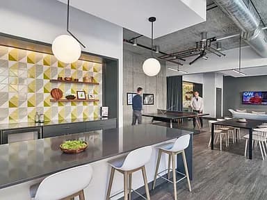 Open-concept lounge with black counter, white bar stools, geometric yellow and white accent wall, pendant lighting, and ping pong table in background.