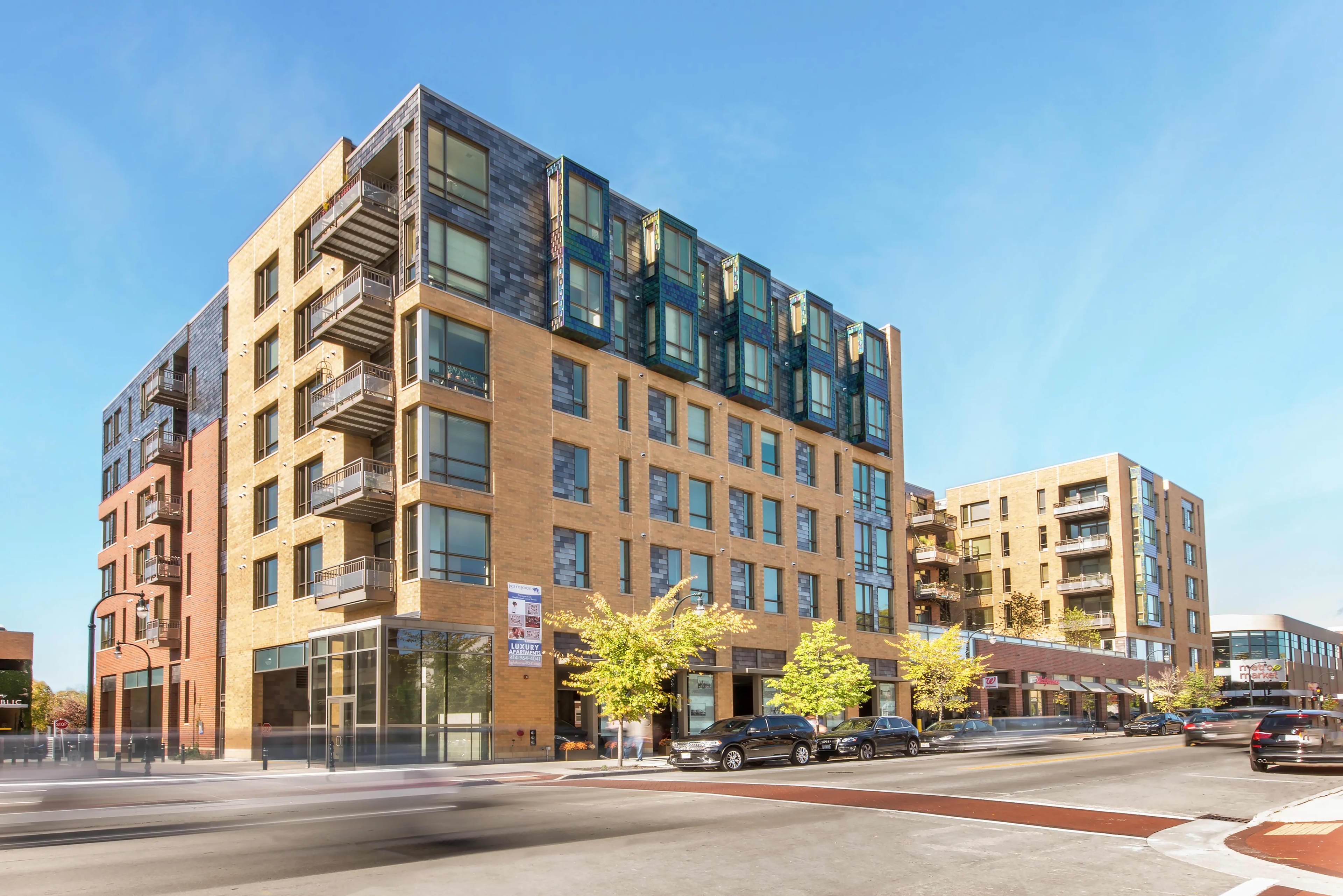 Modern apartment building with brick and glass exterior, metal balconies, ground-floor retail space, and street-level trees in Shorewood.