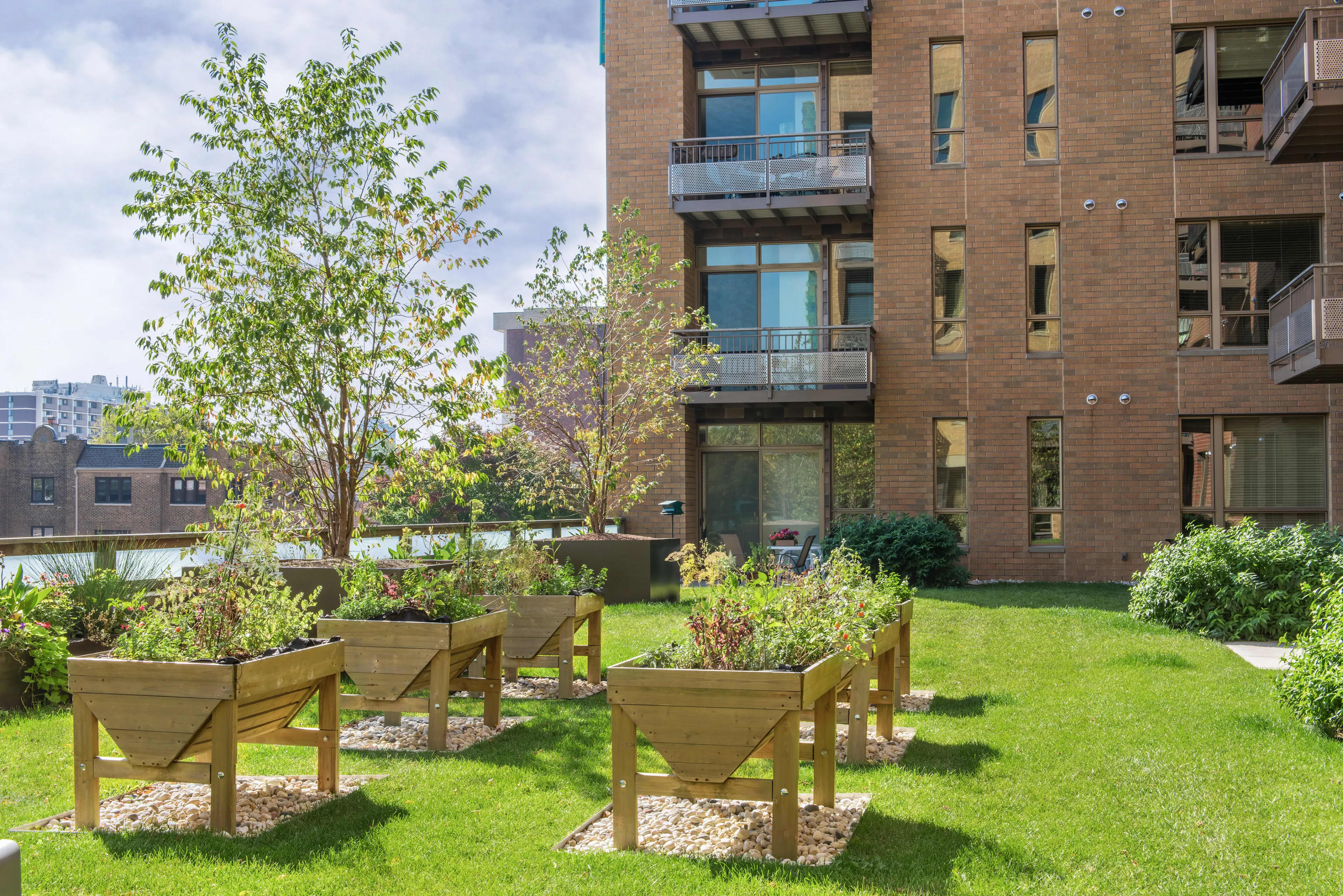 Brick residential building with balconies overlooking a green courtyard with raised wooden planters, mature trees, and water views in the background.
