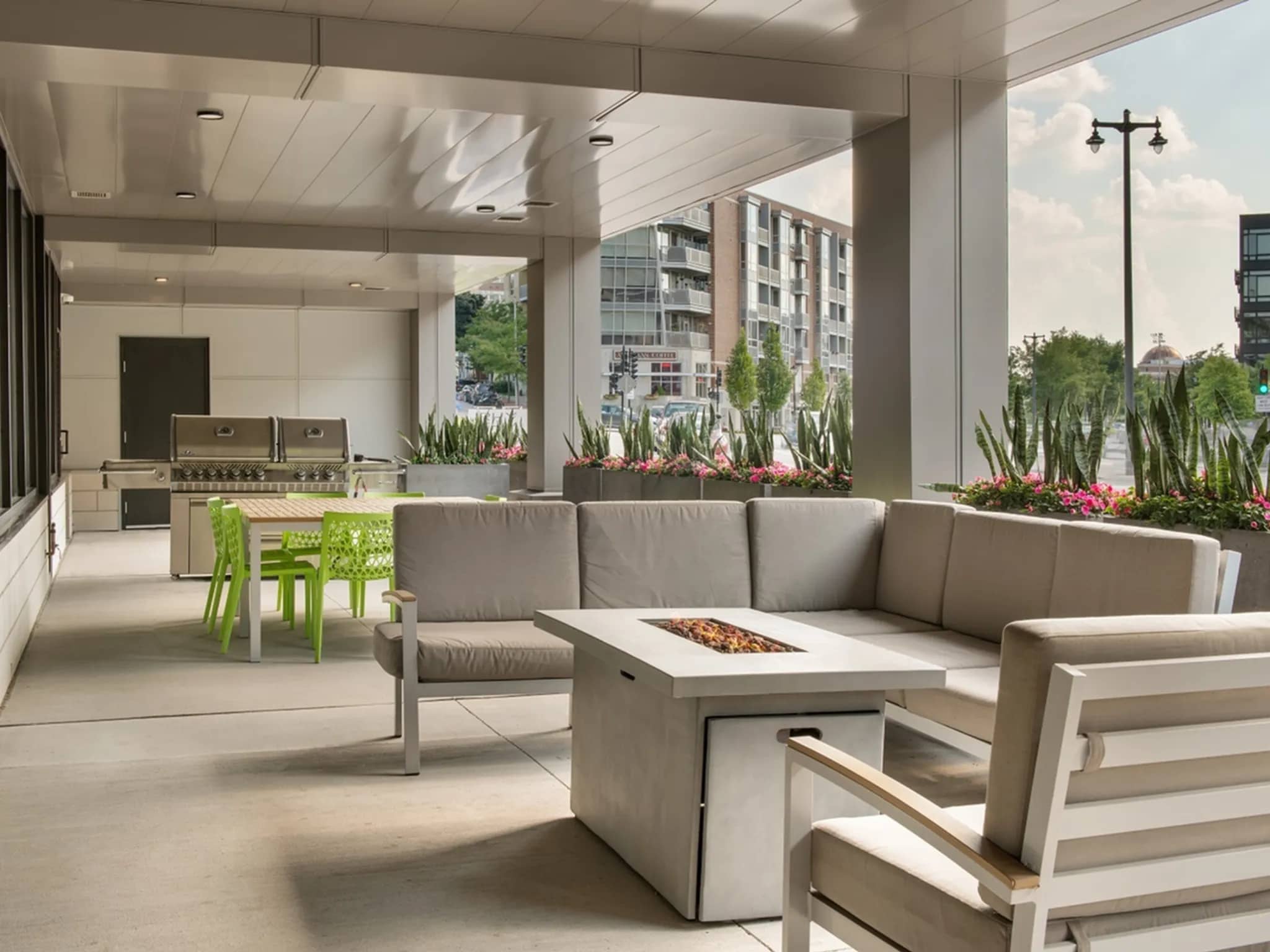 Covered outdoor lounge with L-shaped gray seating, white fireplace table, potted plants, and kitchen area visible through glass doors.