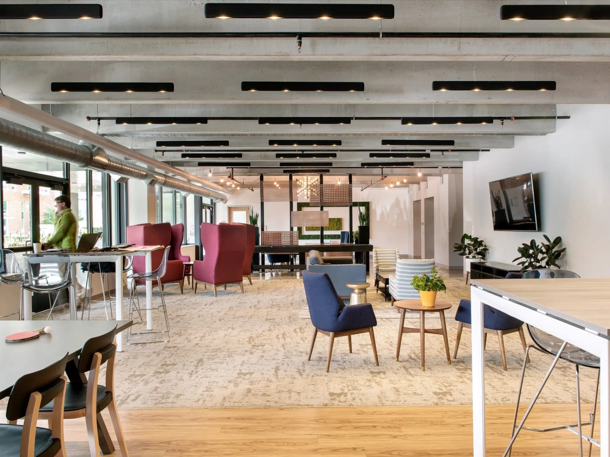 Open common area with burgundy and blue chairs, wooden tables, linear ceiling lights, and potted plants throughout.