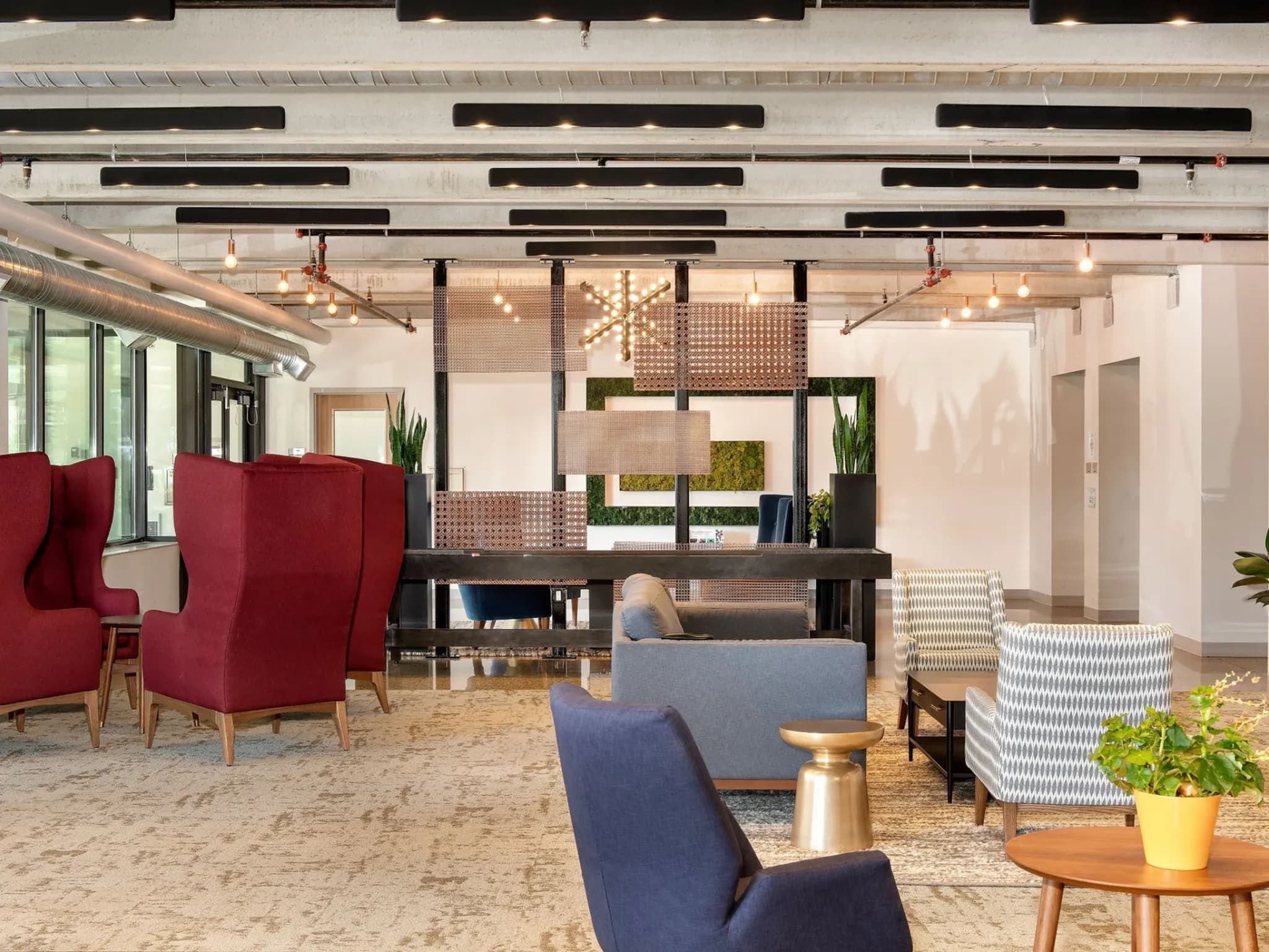 Open-concept common area with red wingback chairs, blue upholstered seating, geometric wood wall dividers, pendant lights, and potted plants.