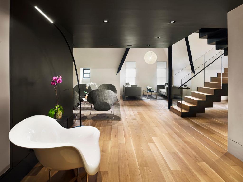 Modern loft interior with white sculptural chair, wooden staircase with black railings, gray seating area, and high ceilings with track lighting.