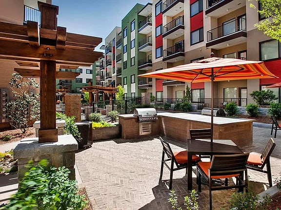 Outdoor courtyard with dining tables, orange umbrellas, wooden pergola, and multi-story apartment buildings in background.