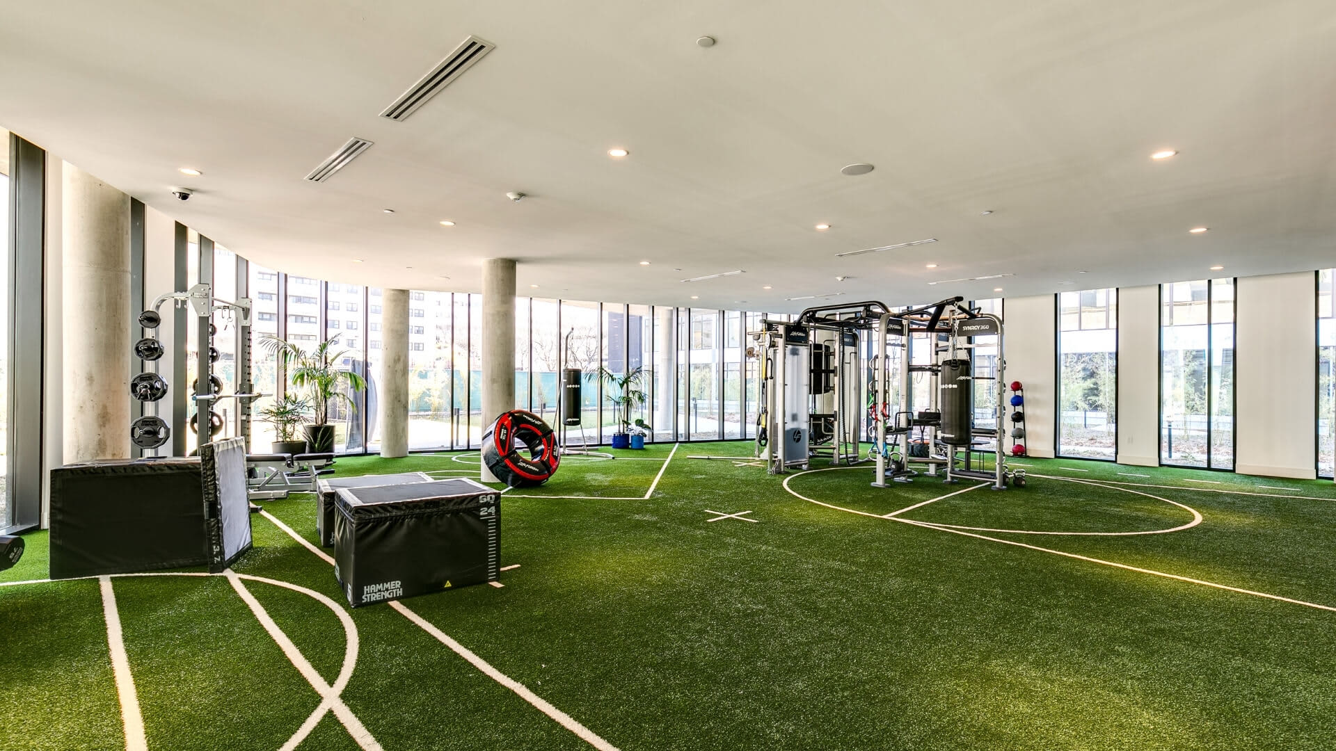 Modern fitness facility with artificial turf flooring, exercise machines, weight storage, and expansive windows with city views.
