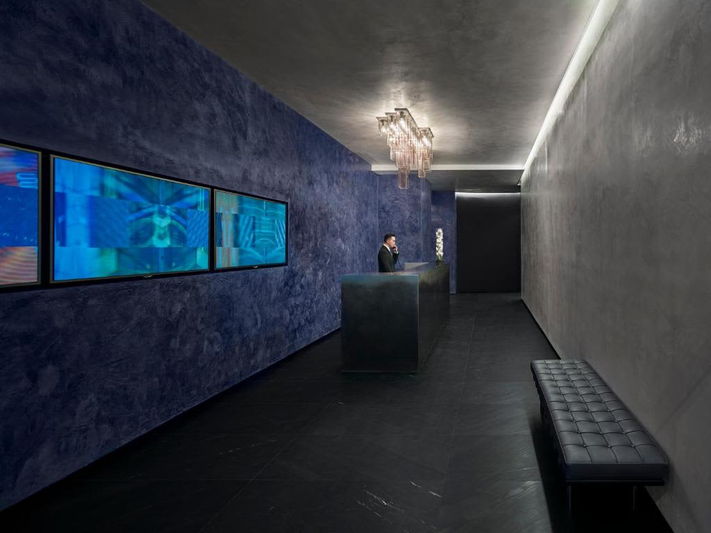 Modern lobby with dual blue-lit digital screens, textured blue-gray walls, crystal chandelier, and black seating bench.