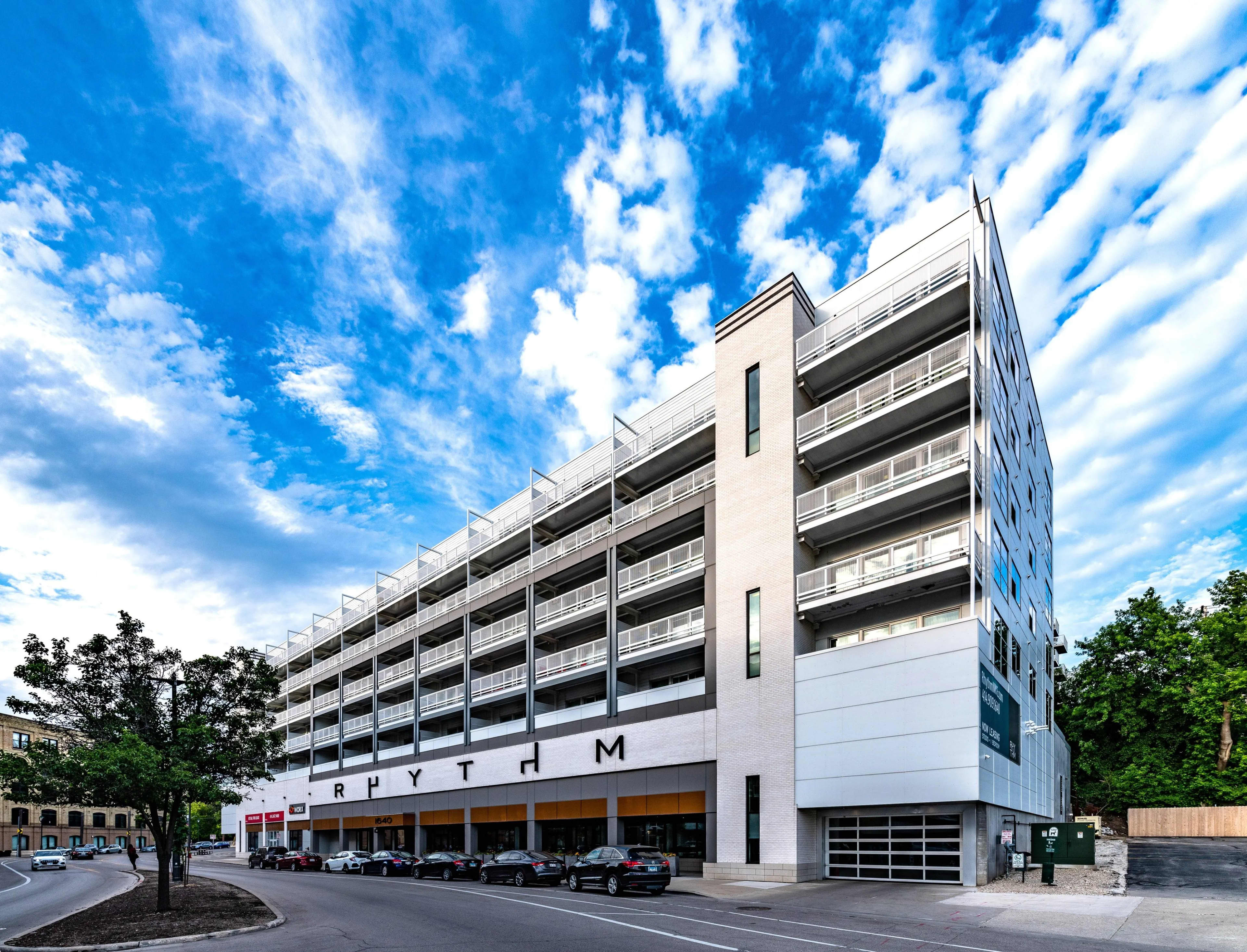 Modern multi-story residential building with white and blue facade, ground-level retail spaces, and parked cars beneath clear blue sky.