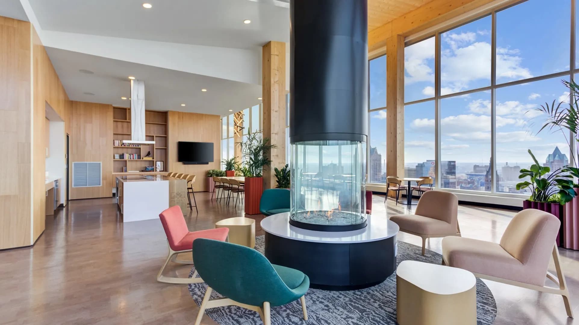 Open living room with central cylindrical fireplace, colorful mid-century chairs, wood cabinetry, and floor-to-ceiling windows overlooking Milwaukee skyline.