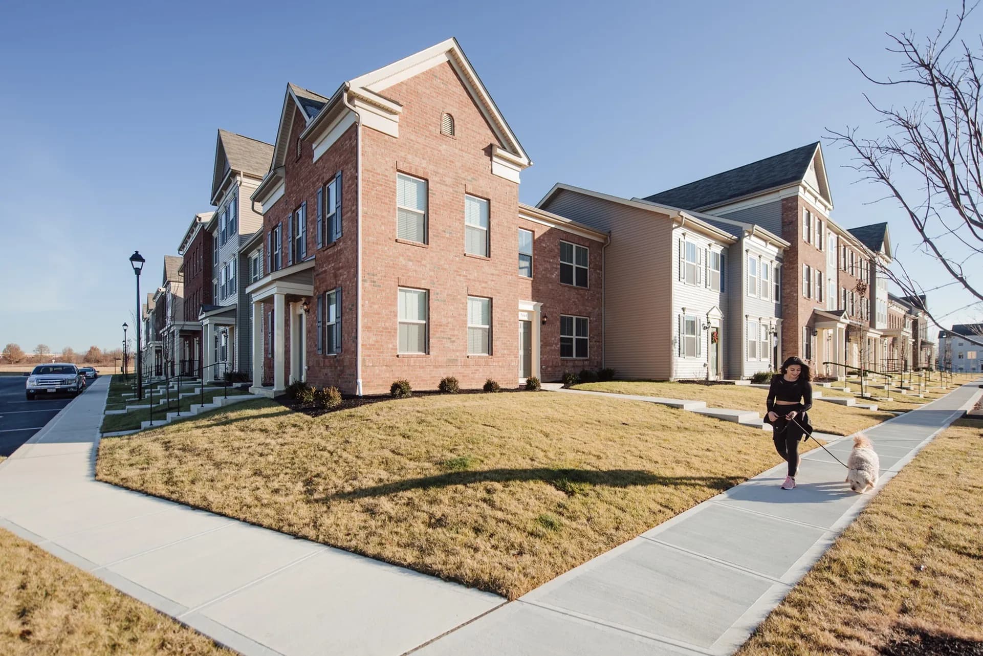 Row of brick and tan siding townhomes with gabled roofs, front lawns, and concrete sidewalks in a residential community.