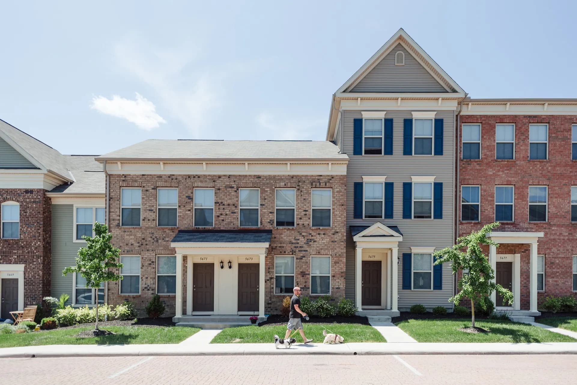 Row of three-story townhomes with brick and siding exteriors, dark shutters, and front porches along a tree-lined street.