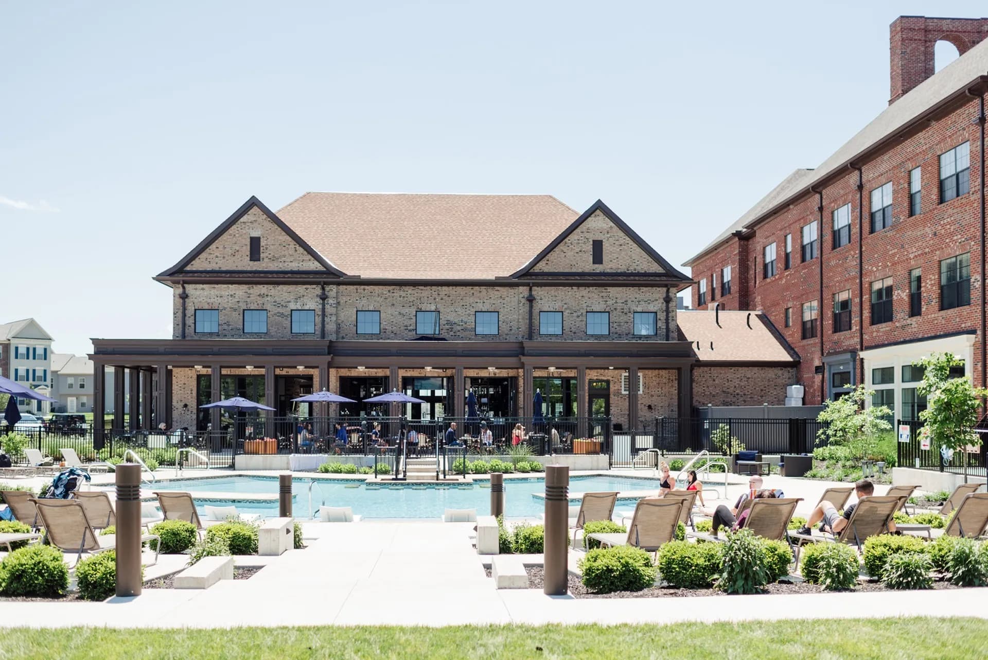 Outdoor pool area with tan lounge chairs, brick clubhouse building with covered patio, umbrellas, and manicured landscaping.