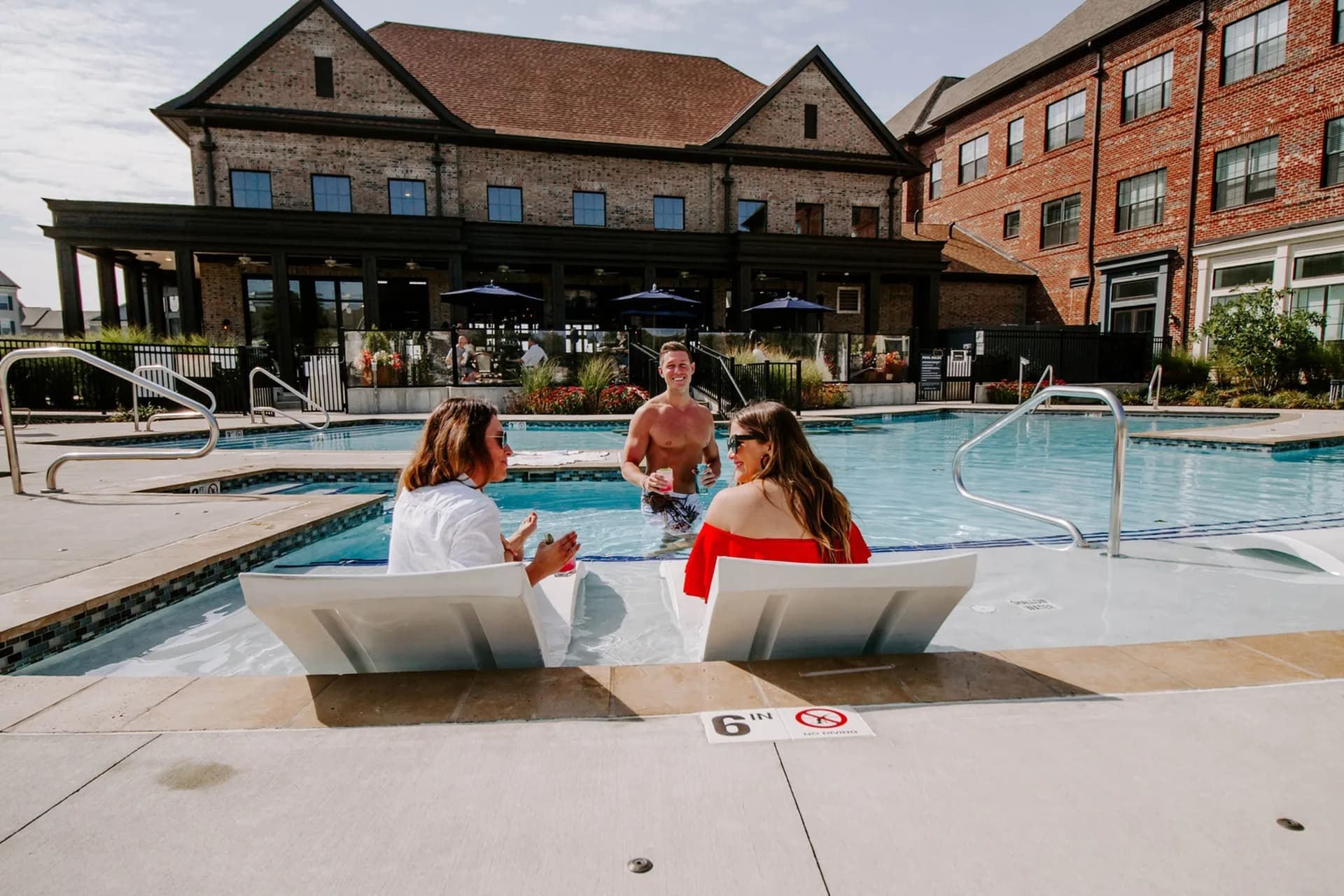 Three people enjoying a resort-style swimming pool with lounge chairs, umbrellas, and a covered pavilion building in the background.