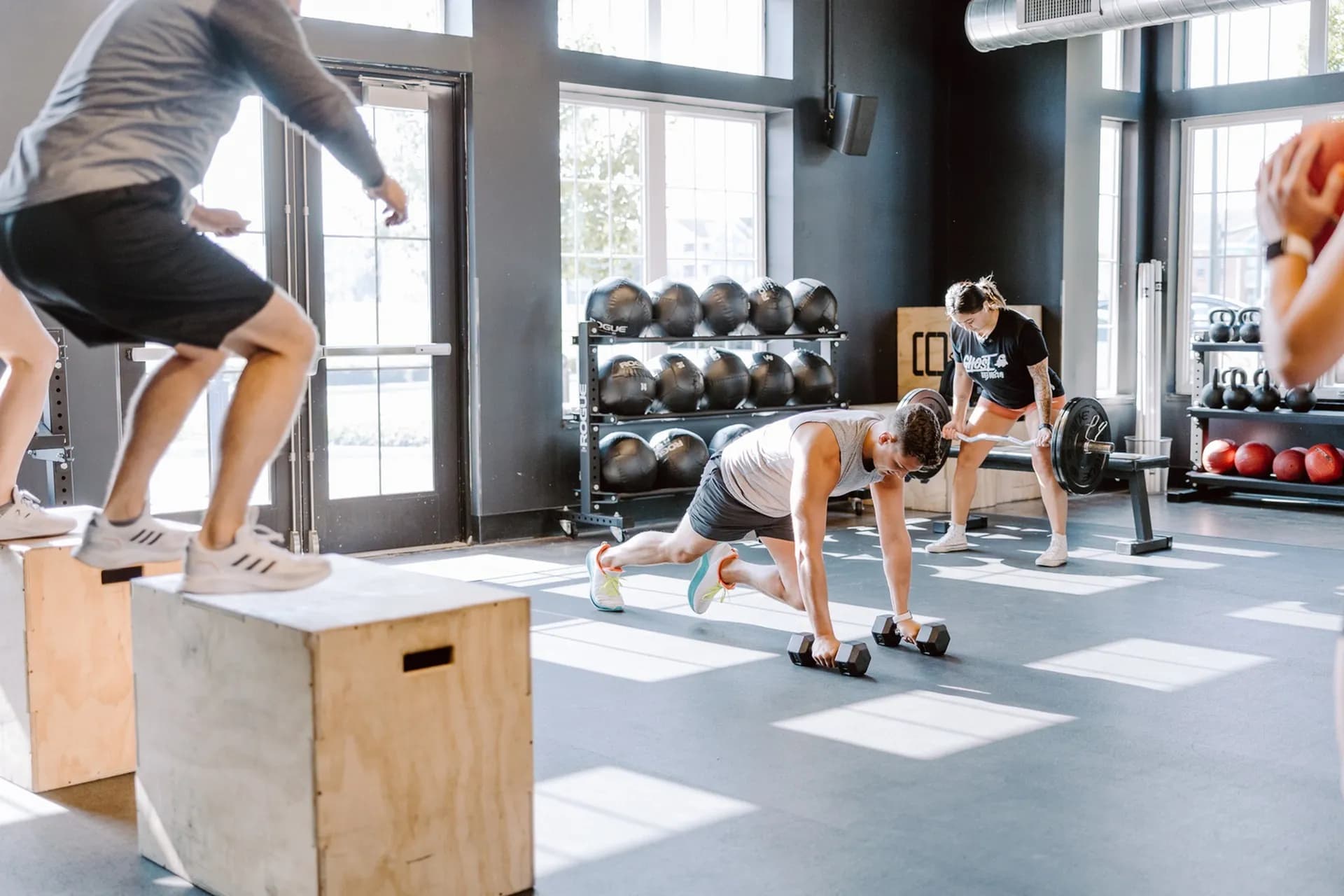 Modern gym with multiple people exercising, shelves of medicine balls, dumbbells, and large windows with natural light.