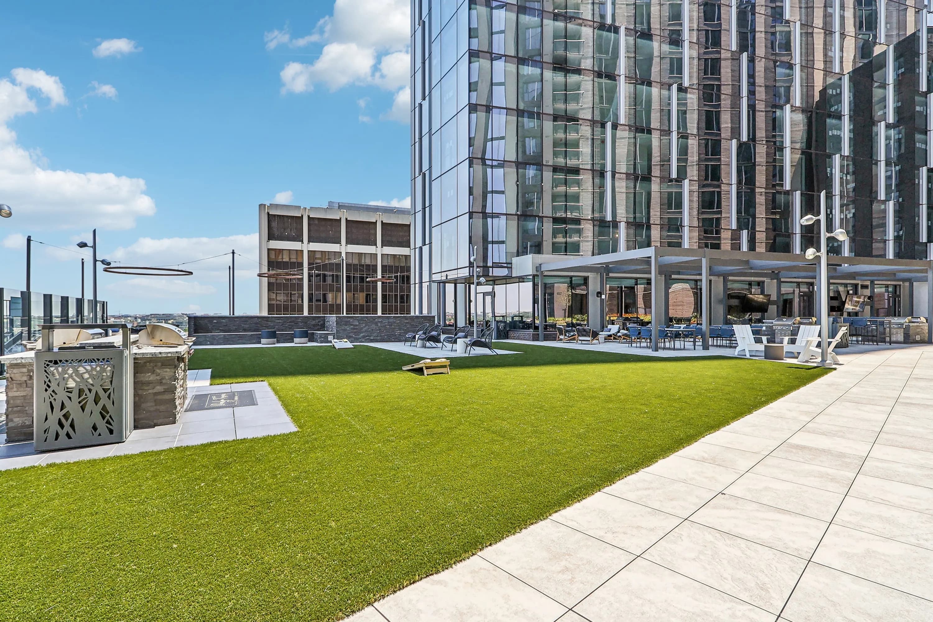 Rooftop terrace with bright green lawn, paved walkway, modern lounge furniture, pergola structure, and surrounding high-rise buildings under blue sky.