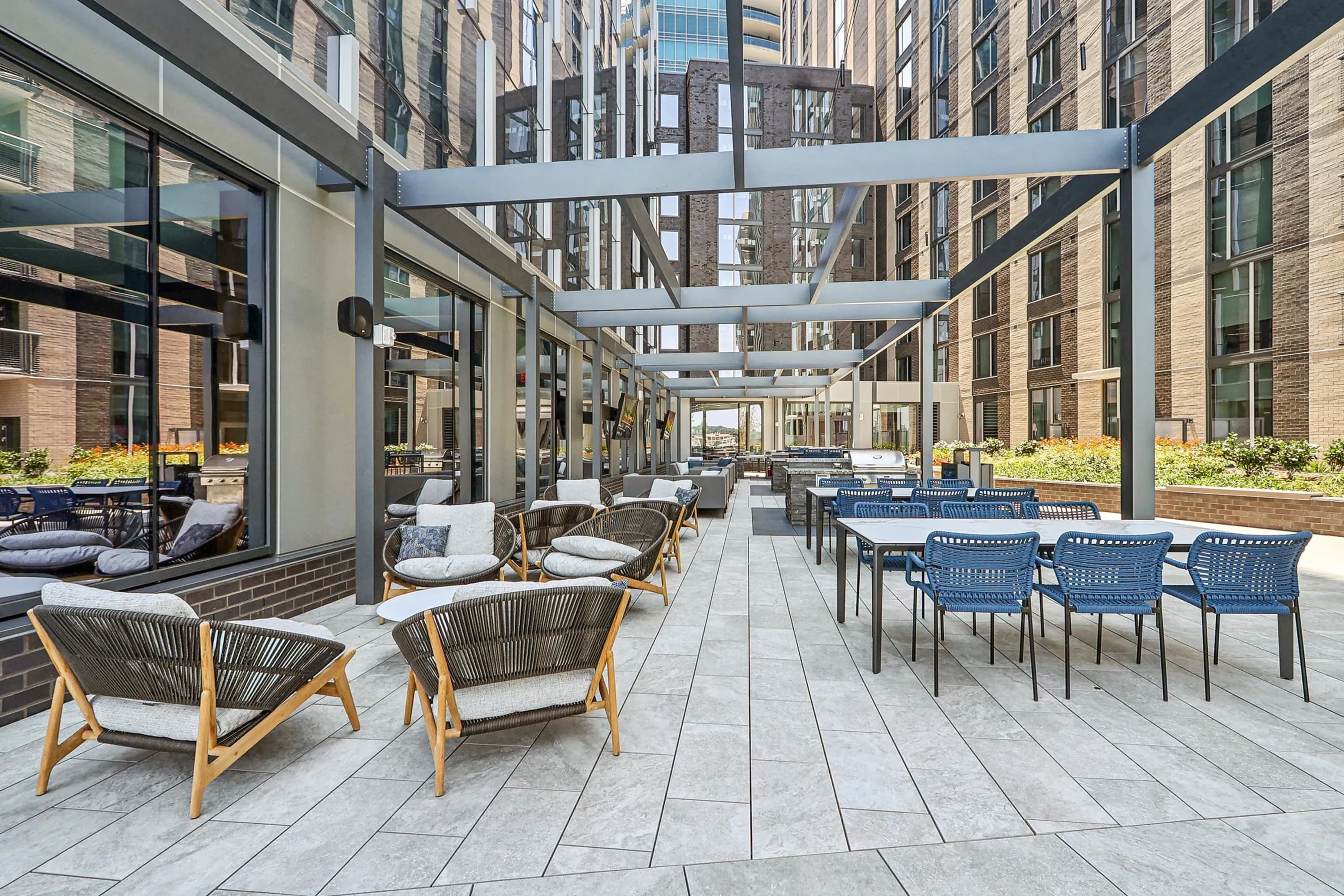 Rooftop terrace with wooden lounge chairs, blue dining chairs, gray pergola, and brick building facades in urban setting.