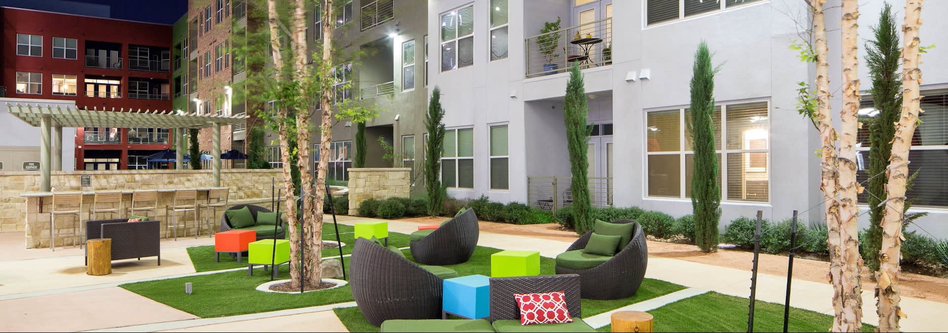 Outdoor courtyard with black woven lounge chairs, colorful accent seating, tall cypress trees, and multi-story residential buildings surrounding manicured lawn areas.