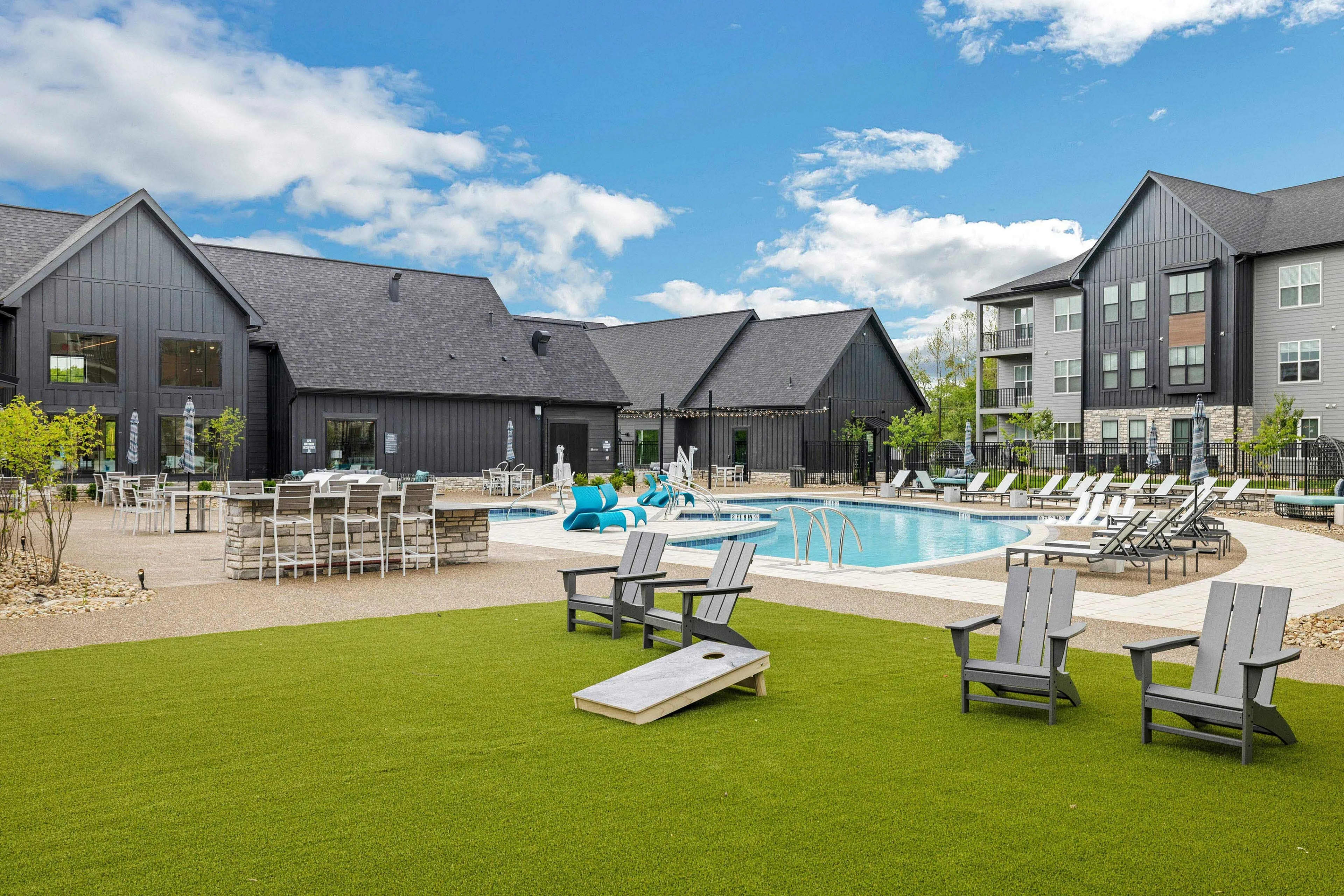 Resort pool with blue water, Adirondack chairs on lawn, cornhole game, and modern black buildings with gray roofs under clear sky.