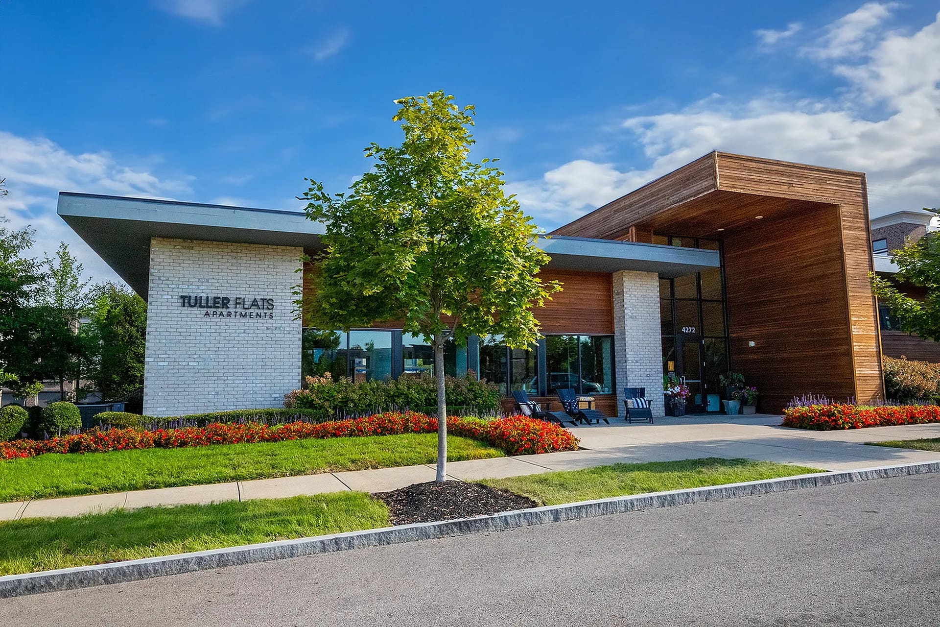 Modern apartment leasing office with white brick facade, wood paneling, large windows, and red flowering shrubs under blue sky.