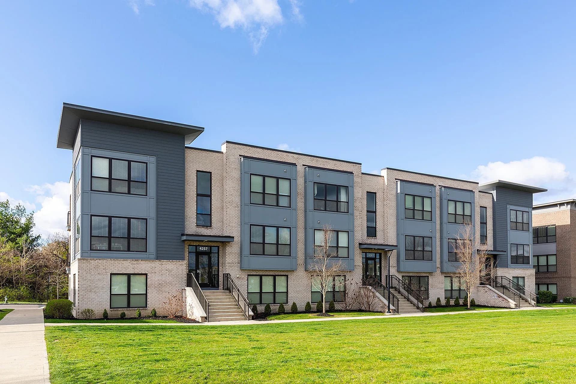 Modern multi-unit residential building with brick and gray siding, large windows, front entrances with stairs, and green lawn.