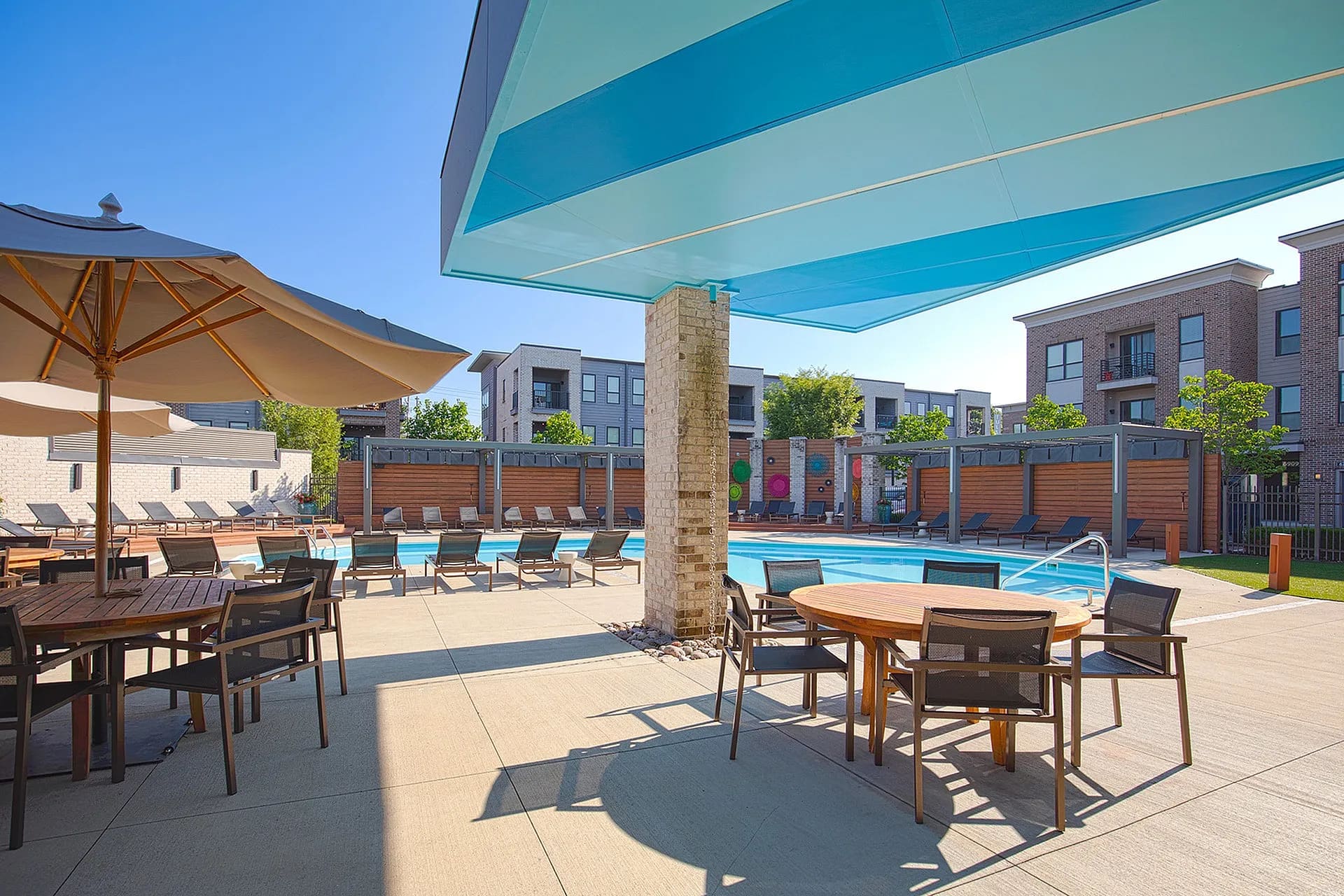 Resort-style pool deck with blue water, lounge chairs, dining tables, umbrellas, and modern residential buildings in background under clear sky.