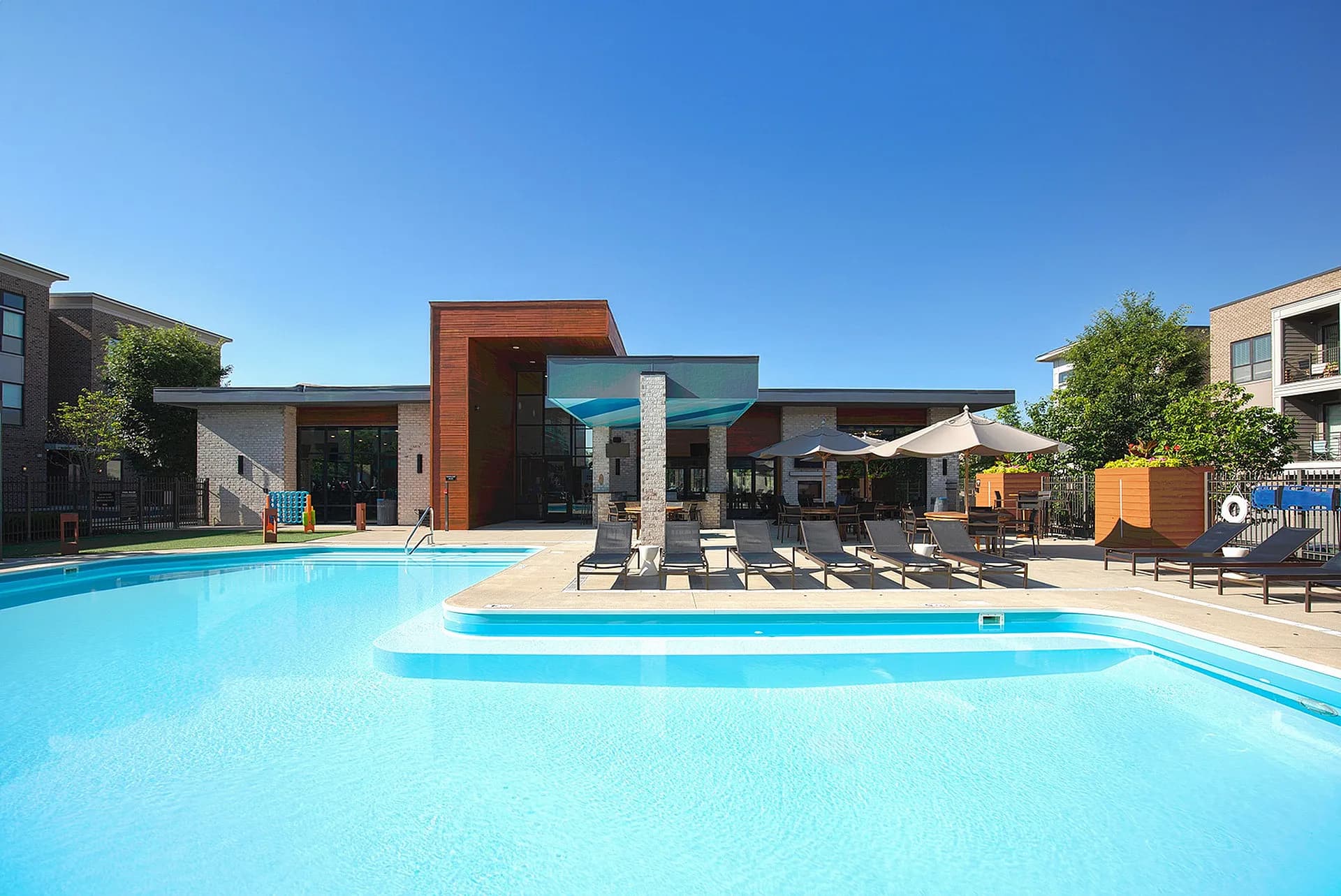 Two bright blue swimming pools with a modern brick and wood clubhouse, lounge chairs, and shade structures in a residential community.