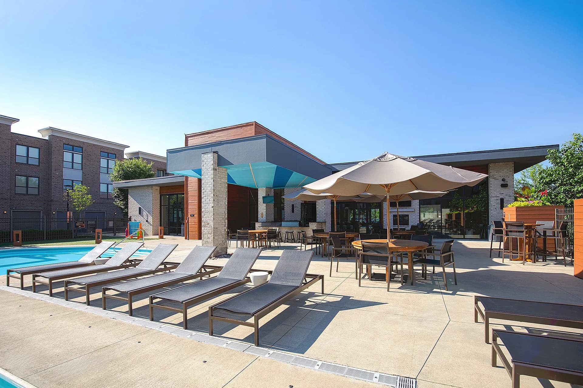 Pool deck with lounge chairs, umbrellas, dining tables, and modern clubhouse building under clear sky.
