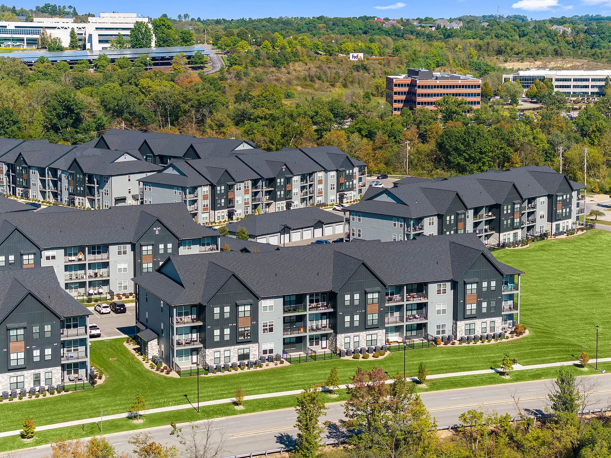 Aerial view of multi-story apartment complex with dark roofs, mixed dark and light facades, manicured lawns, and surrounding wooded area.