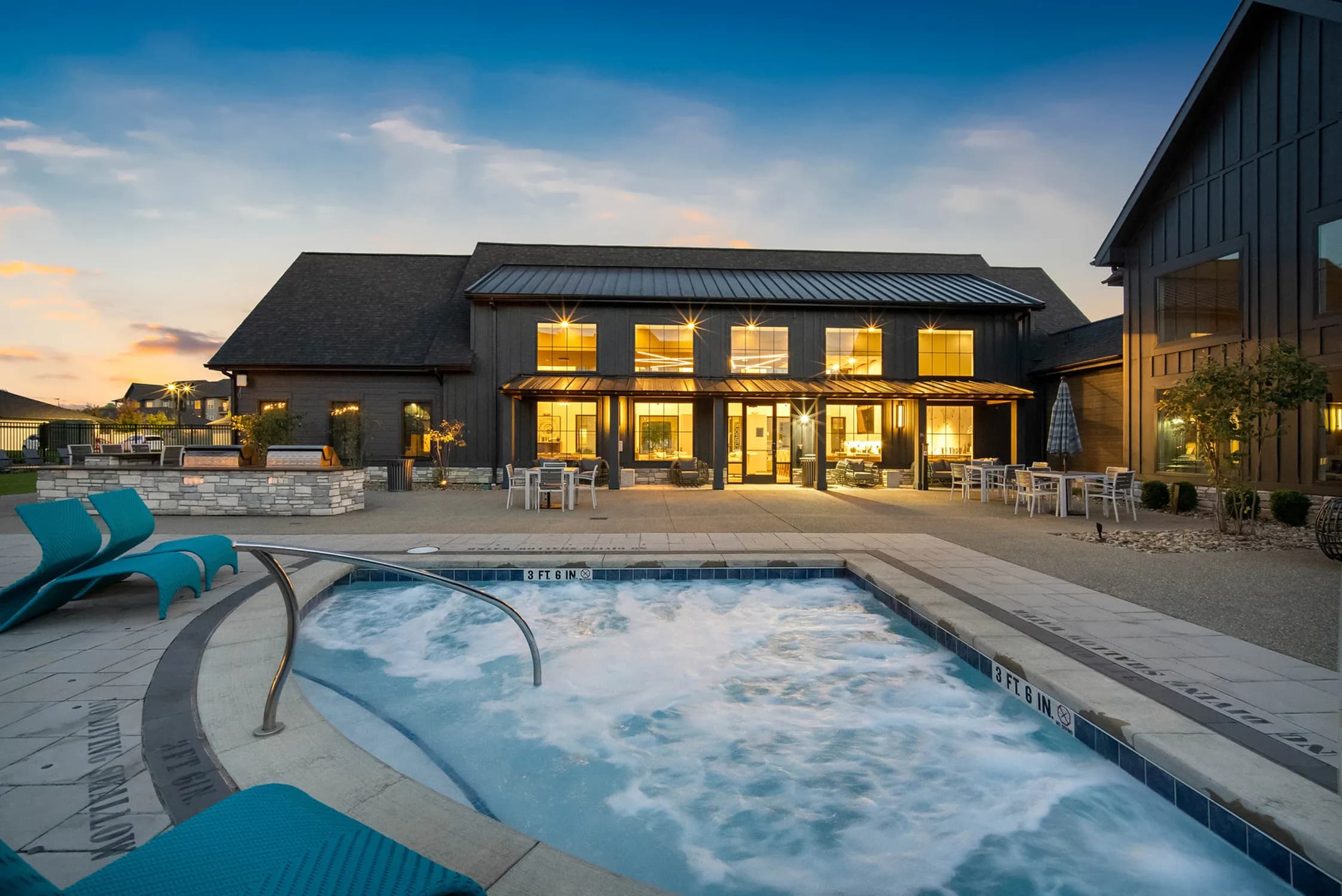 Heated swimming pool and spa in foreground with turquoise lounge chairs, modern black farmhouse with lit windows and covered patio beyond, stone fire pit area to left.