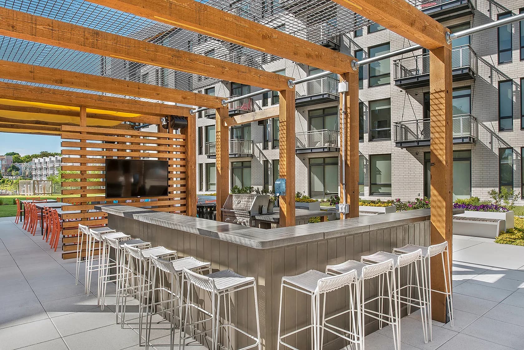 Outdoor bar and lounge area with wooden pergola, gray countertop, white bar stools, TV screen, and residential building in background.