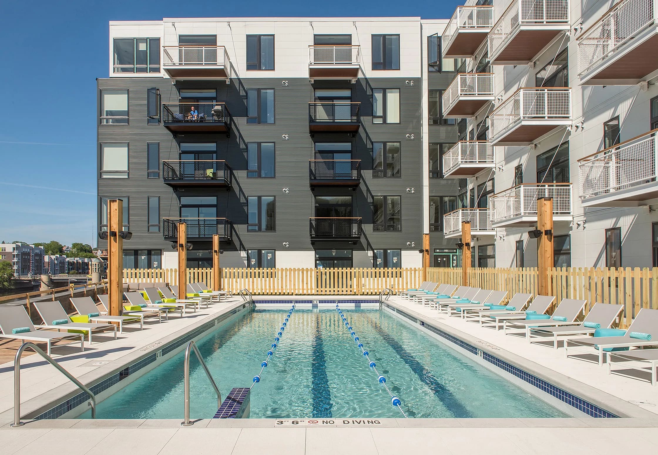 Lap pool with blue lane dividers, lounge chairs on both sides, wooden fence, and multi-story residential buildings under clear sky.