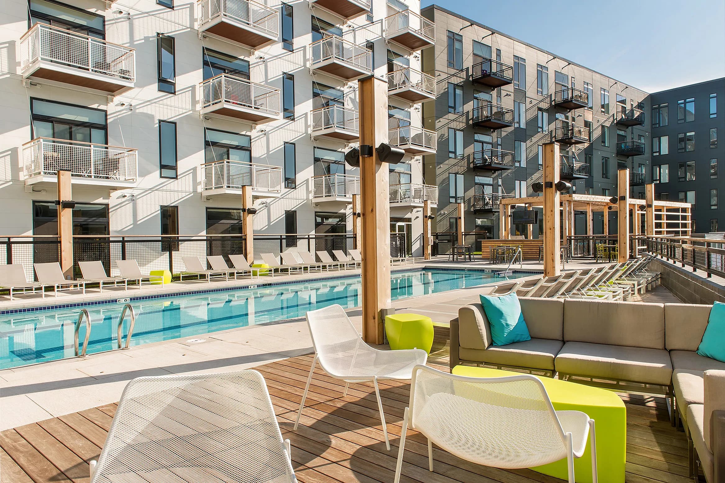Resort-style pool area with lounge chairs, wooden deck, and multi-story apartment building with balconies in background.