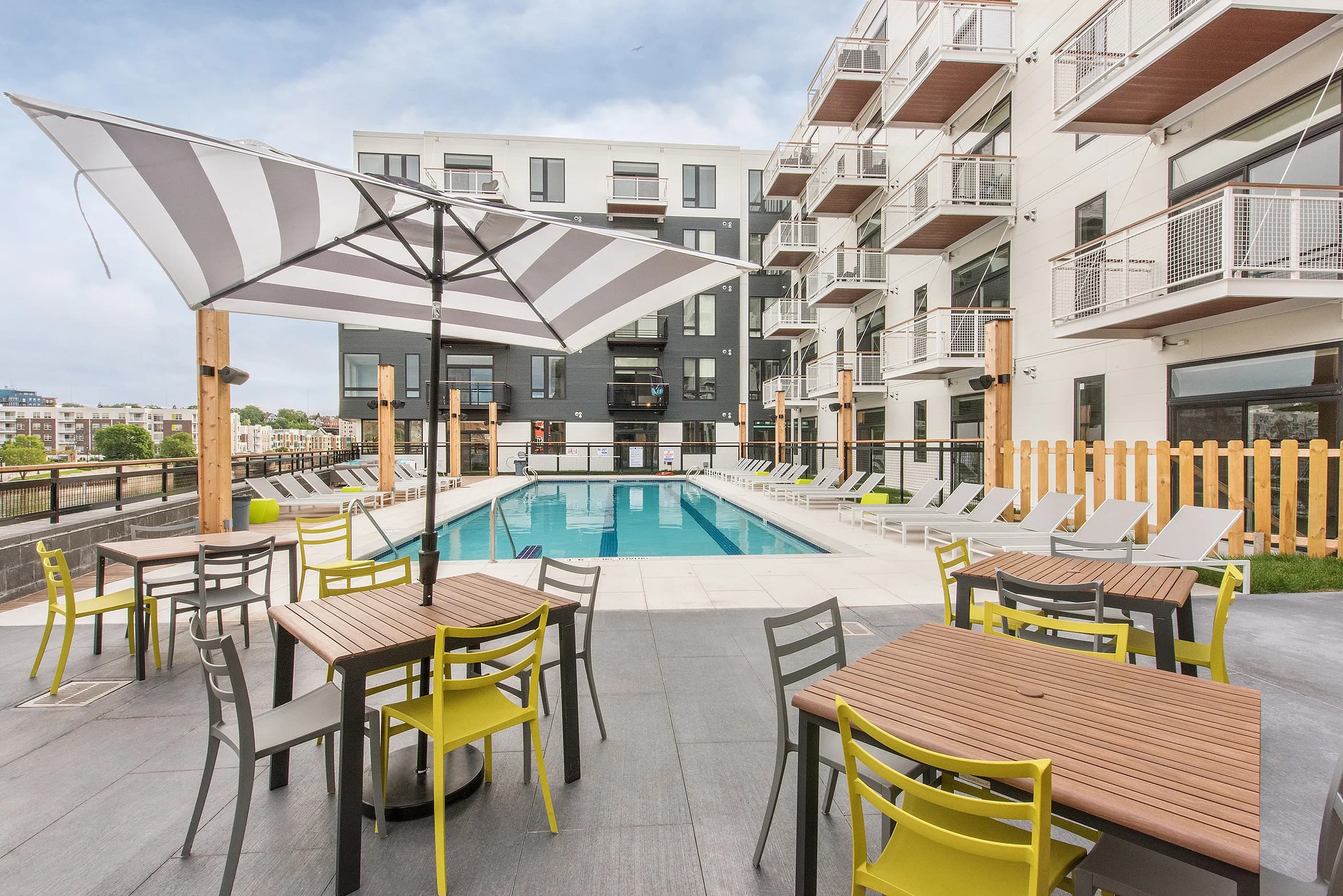 Outdoor pool deck with turquoise swimming pool, yellow and gray chairs, striped umbrella, wooden tables, and multi-story residential building.