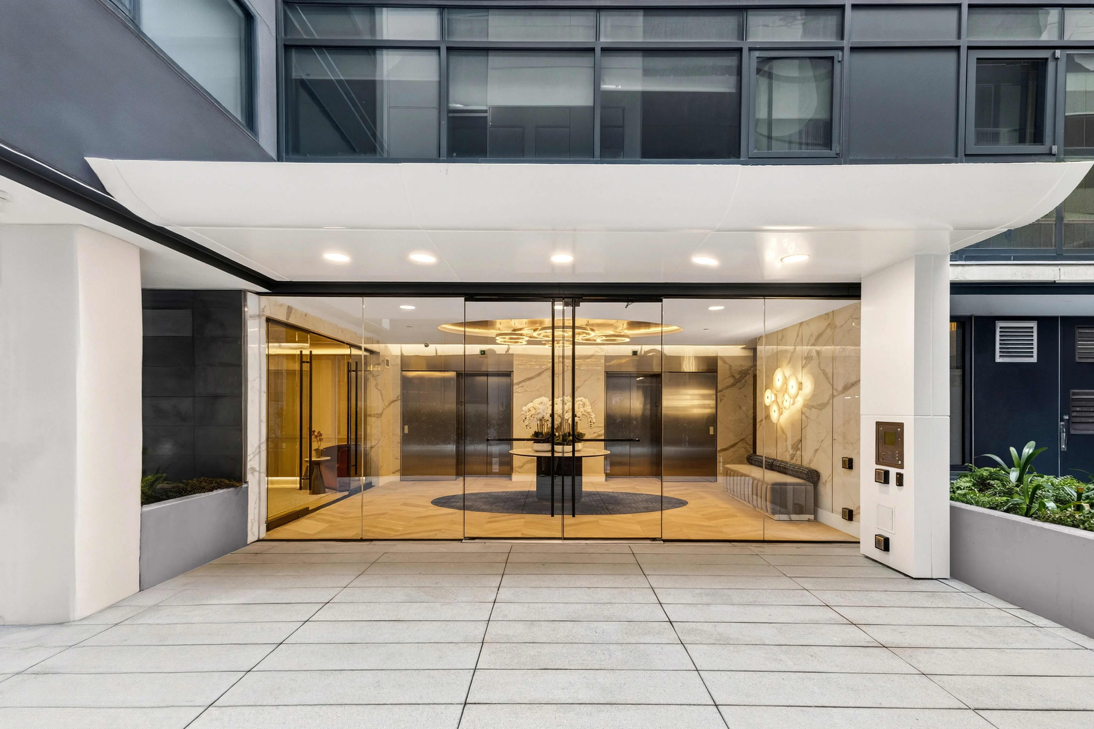 Modern building lobby with large glass doors, polished wood interior, recessed lighting, and light gray tile flooring.