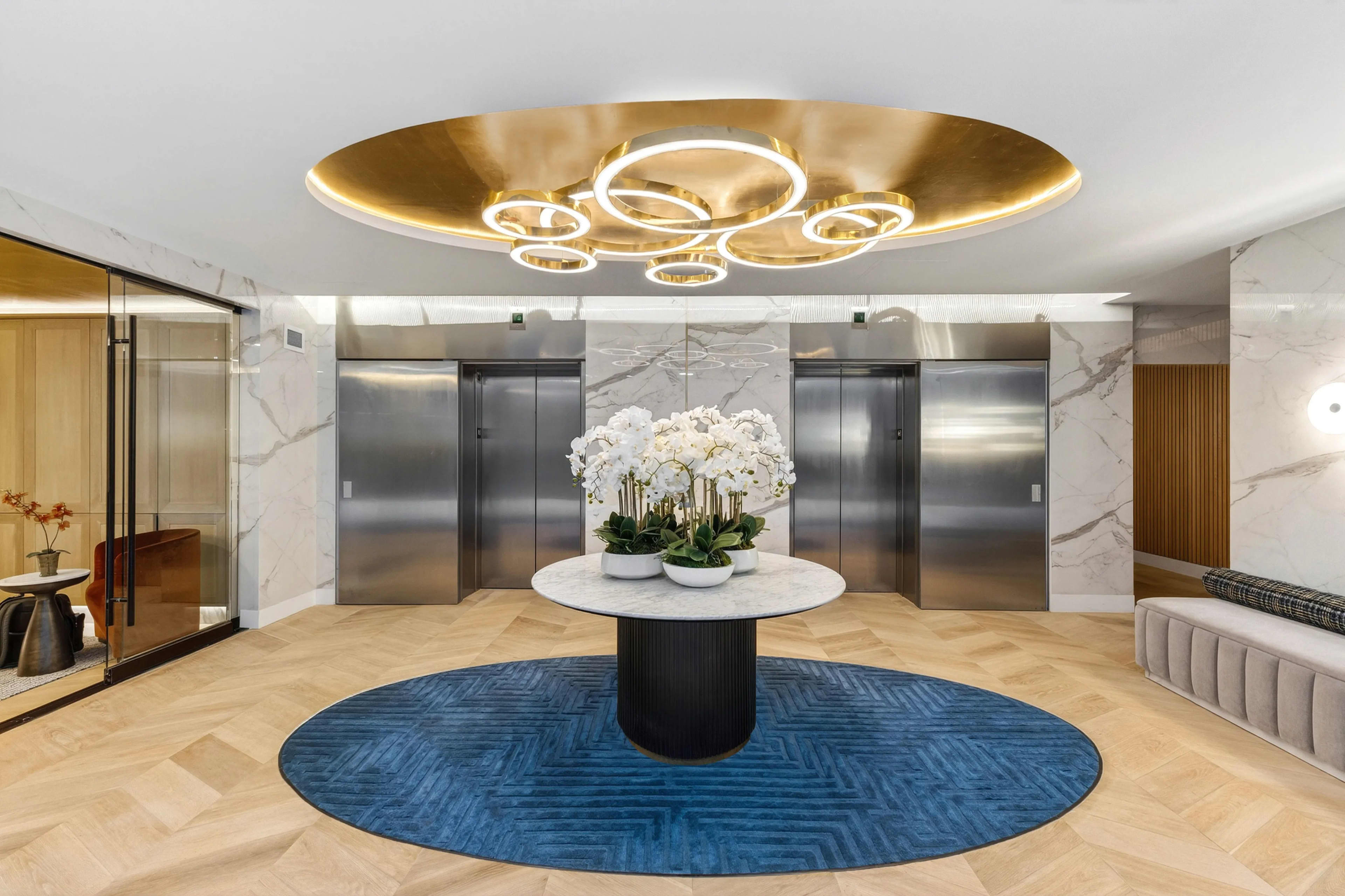 Modern lobby with stainless steel elevator doors, marble walls, circular blue rug, and brass ring ceiling fixture with white orchid centerpiece.