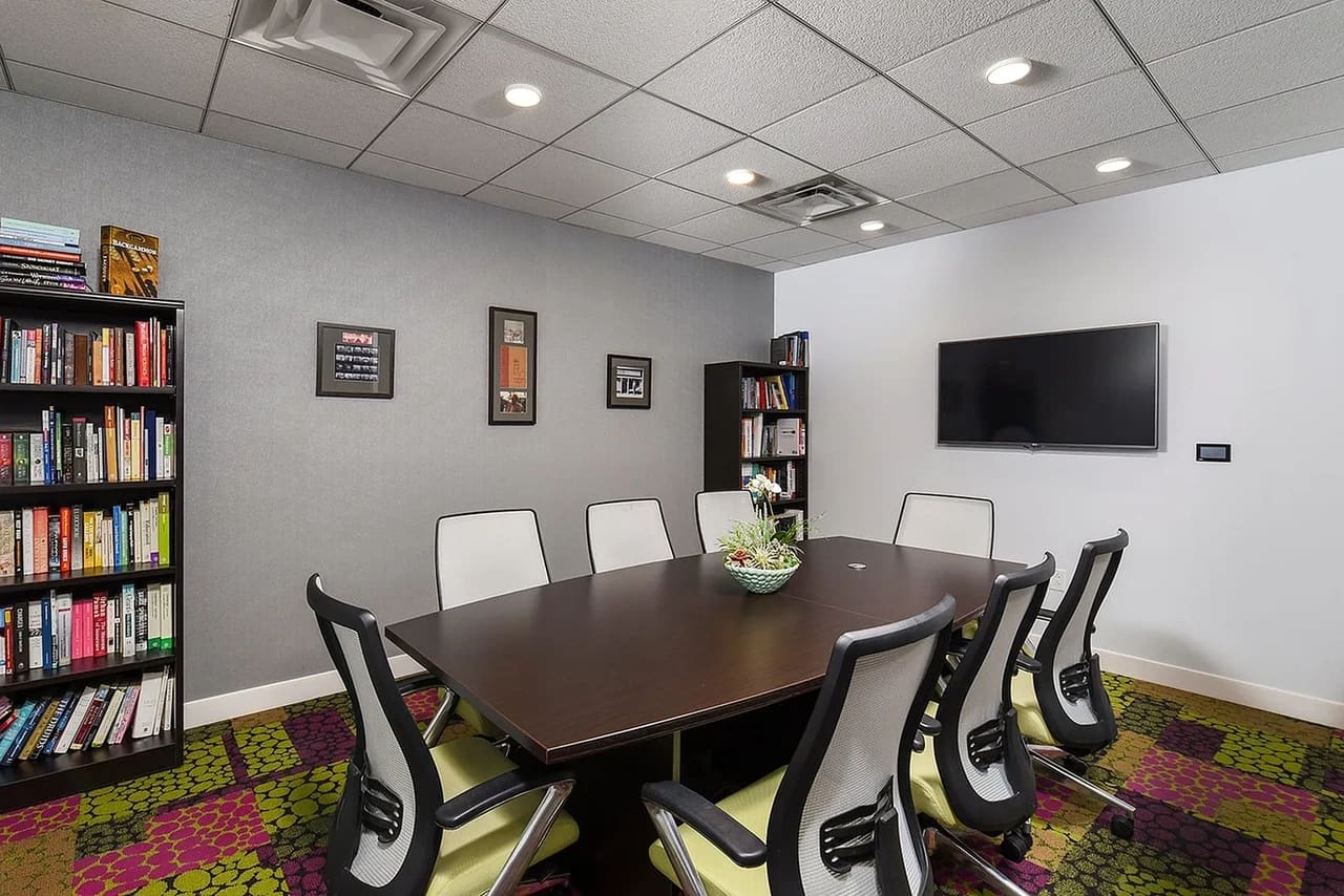 Conference room with dark wood table, six ergonomic chairs, colorful geometric carpet, wall-mounted TV, and floor-to-ceiling bookshelves.