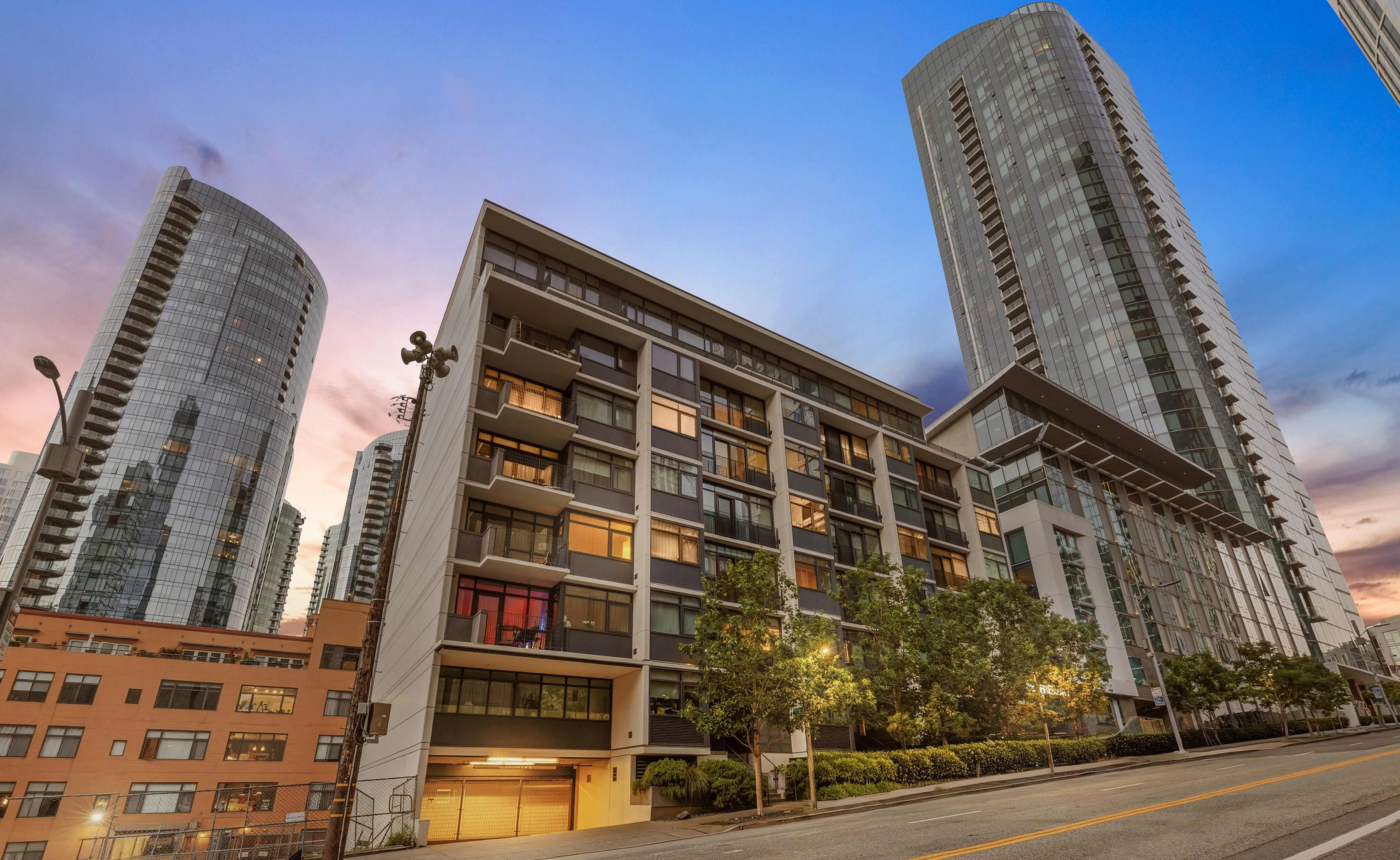 Mid-rise residential building with concrete and glass facade, flanked by tall glass towers, trees, and street-level parking at dusk.