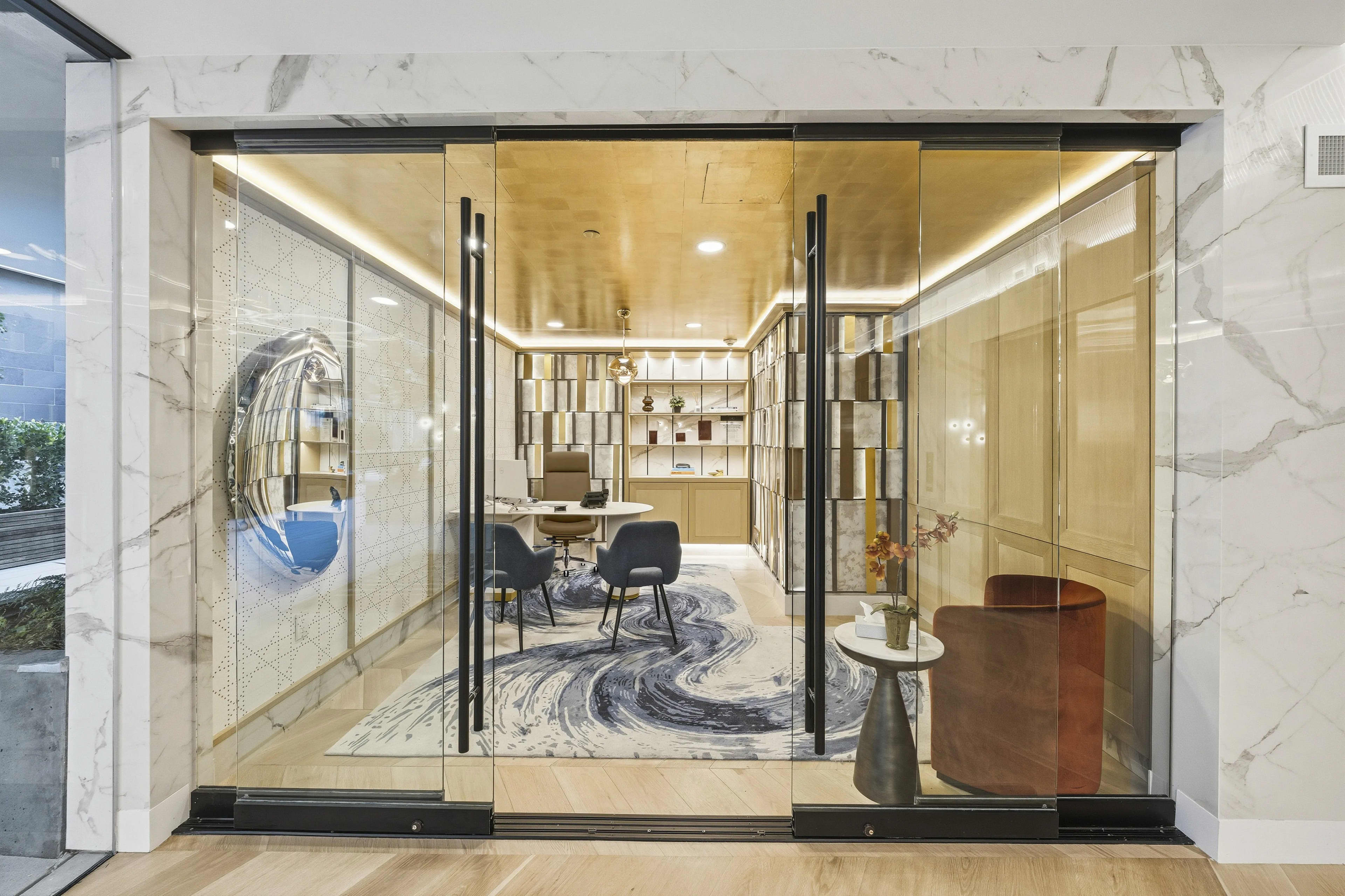 Studio apartment with glass partition walls, warm wood ceiling, patterned area rug, black chairs, and built-in shelving visible through open glass doors.