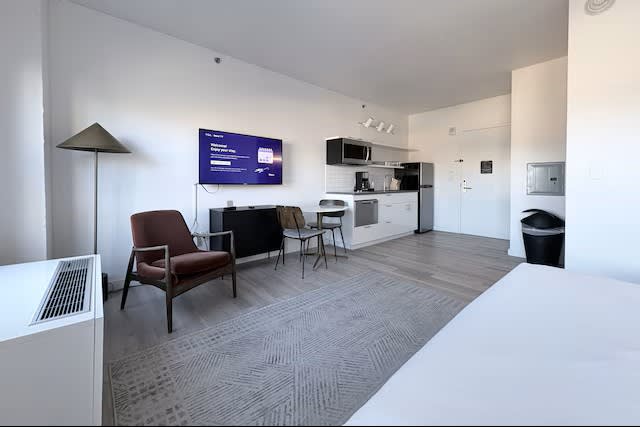 Studio apartment with brown mid-century chair, kitchenette with white cabinetry, desk area, and gray patterned flooring.