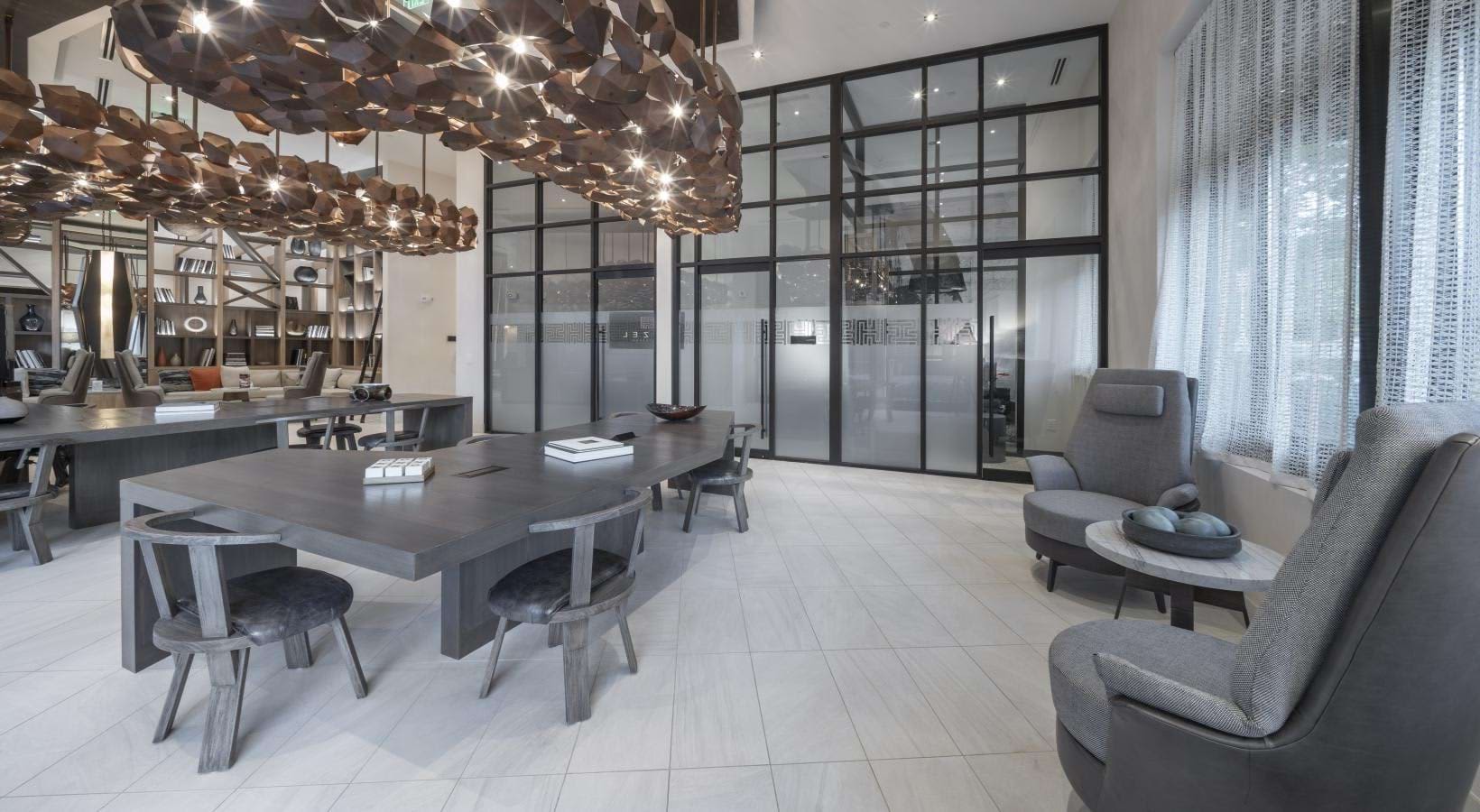 Open-concept common area with gray dining table, gray lounge chairs, sculptural ceiling fixture, and floor-to-ceiling windows with sheer curtains.